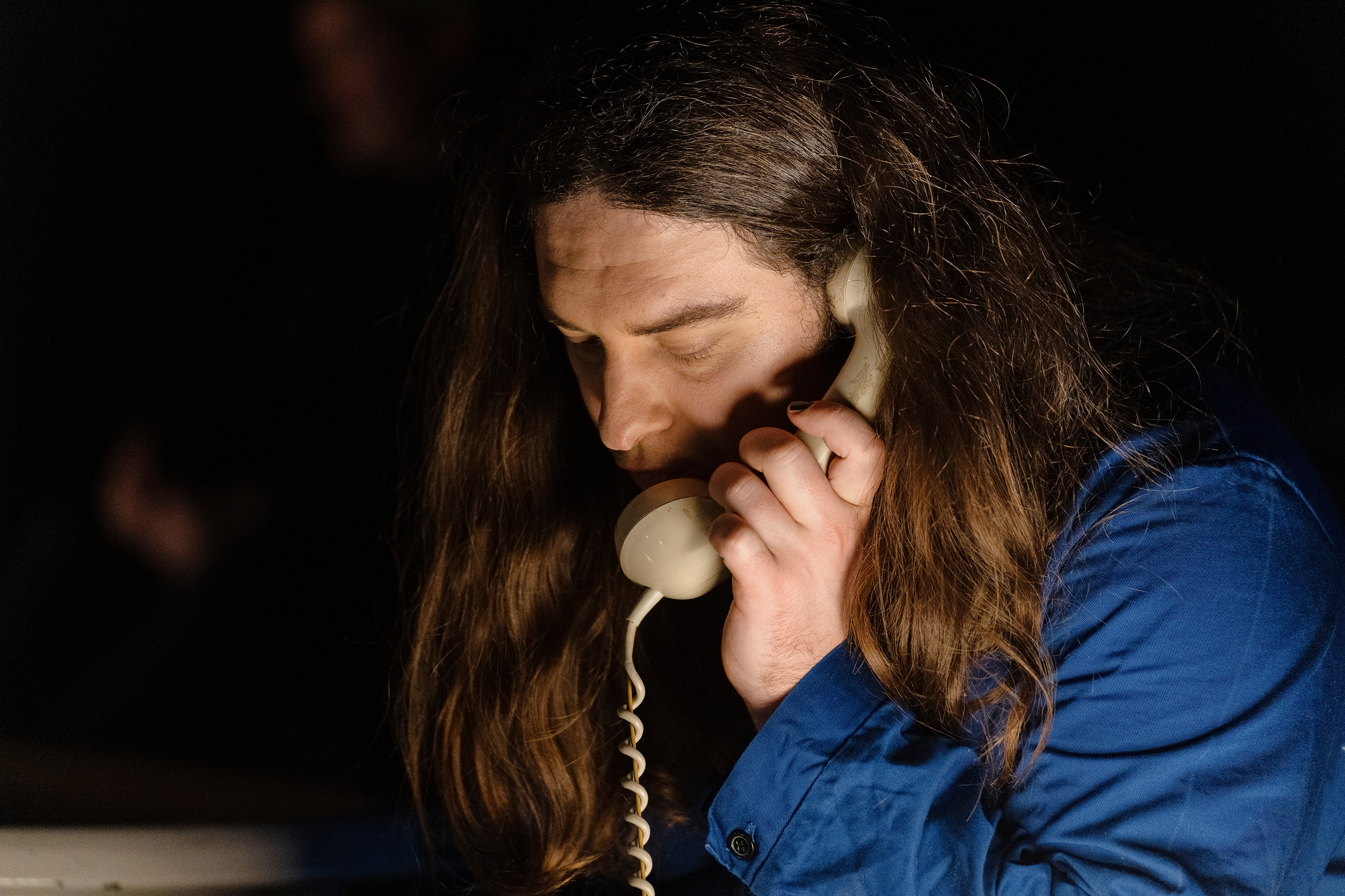 Auf dem Bild ist der Kopf einer weißen Person mit langen dunkel braunen Haaren zu sehen. Sie trägt einen Blauen Overall und hat einen beigen Telefonhörer in der linken Hand. Der Hintergrund ist dunkel.